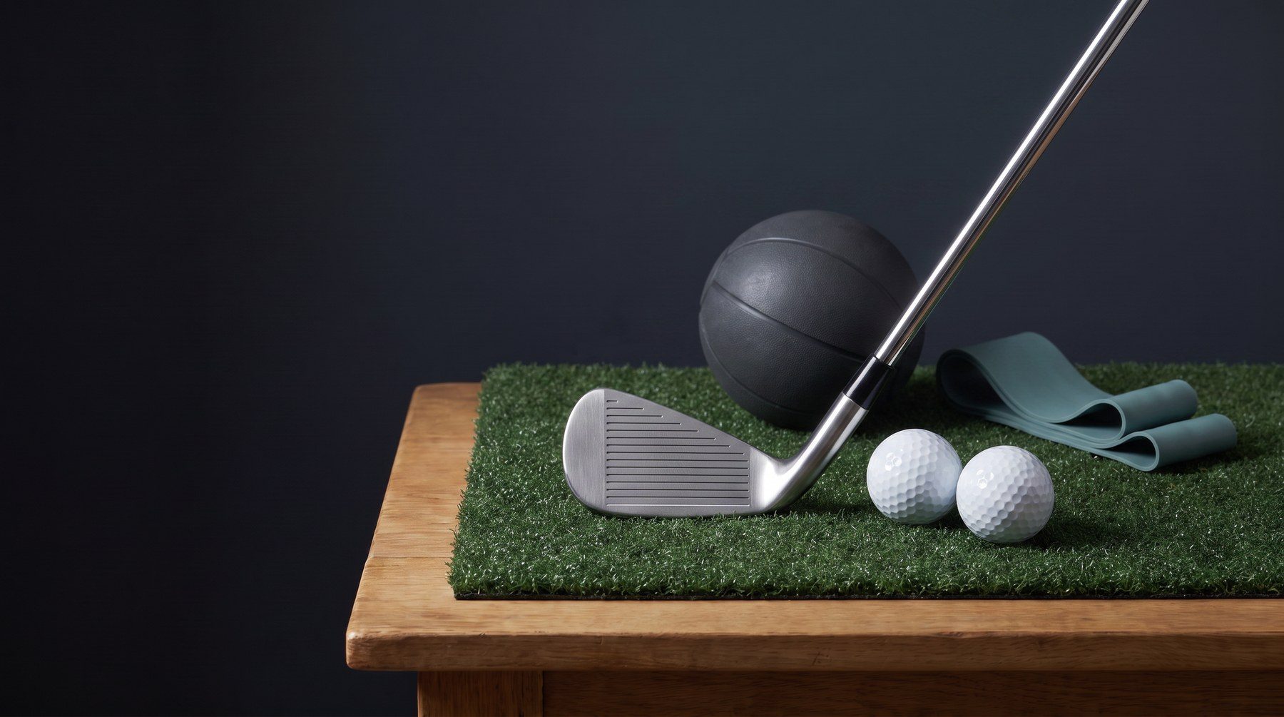 Conceptual rendering of a golf iron and golf balls on practice turf beside a medicine ball and teal resistance band, illustrating golf strength and mobility training