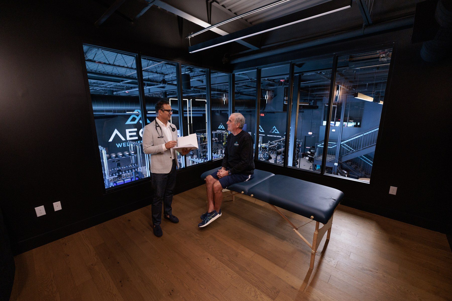A consultation beside a treatment table overlooking the training floor at Aeon Wellness Club
